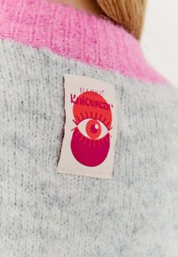 Sweter z dzianiny o szarym korpusie i różowym ściągaczu. Posiada metkę z tkaniny z grafiką oka i tekstem "NAKGKO Knitwear".