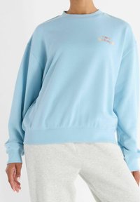 Person wearing a light blue New Balance sweatshirt with orange logo on chest and light gray sweatpants, standing with relaxed posture.