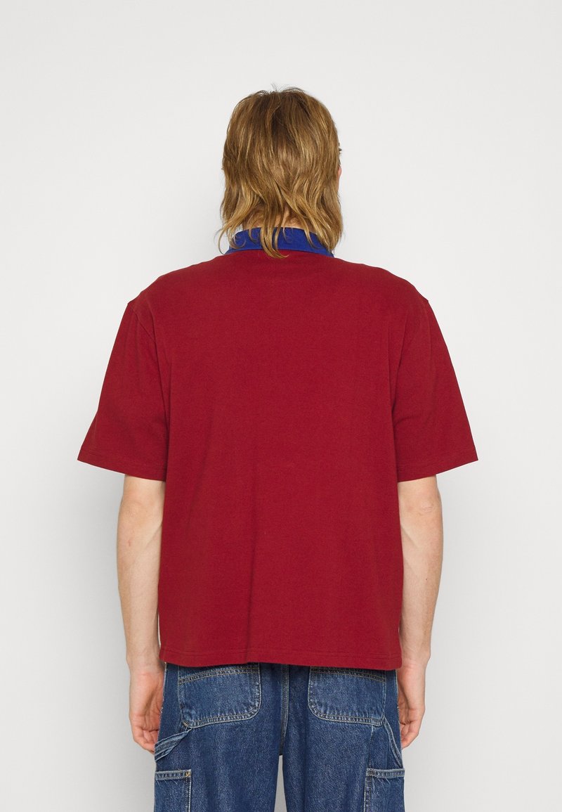 Personne aux cheveux mi-longs portant une chemise rouge à manches courtes et un jean bleu, vue de dos sur un fond blanc uni.