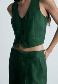 Veste verte ajustée et courte avec un décolleté en V, deux boutons et des rayures verticales blanches. Associée à un pantalon taille haute assorti.