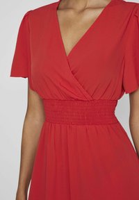 Červené šaty s krátkým rukávem, nařaseným pasem a splývavou sukní. Vyrobené z lehkého materiálu s hladkou texturou.