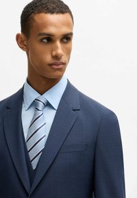 Navy blue suit with a fine check pattern, paired with a light blue shirt and a striped tie in blue and silver tones. Smooth fabric texture.