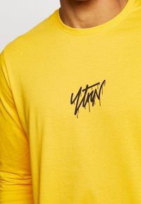 Langärmliges gelbes T-Shirt aus Baumwolle, mit einem schwarzen Graffiti-Logo mit Tropfen-Details auf der Vorderseite. Strukturiertes Gewebe.