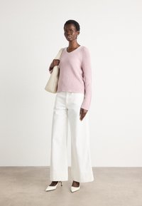Roze geribbelde trui met V-hals, witte wijde broek, en witte puntige hakken. Model heeft een beige tote bag. Lichte achtergrond.