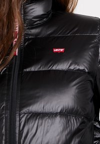 Detailní záběr na osobu, která nosí černou péřovou bundu s logo Levi's na hrudi a červenou podšívku viditelnou u zipu.