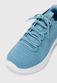 Zapatilla deportiva de color azul claro con parte superior de punto texturizado, cordones planos y suela de goma blanca. Presenta ranuras horizontales sutiles en los laterales.