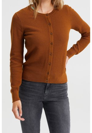 Femme portant un cardigan marron texturé boutonné avec des manches longues et un jean gris foncé, debout avec une main sur la hanche.