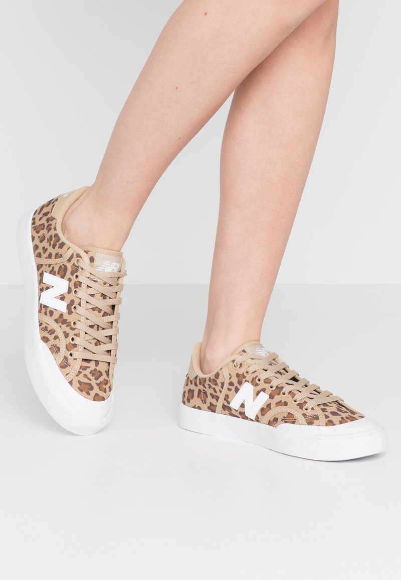 Jambes portant des baskets imprimé léopard beige et marron avec semelles blanches et logo blanc "N", posées sur un fond gris clair.