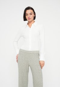 Femme aux cheveux courts et foncés portant une chemise blanche boutonnée et un pantalon gris clair en maille torsadée, sur un fond blanc uni.