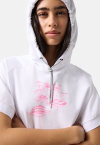 Persona joven con cabello oscuro lleva una sudadera blanca con capucha y mangas remangadas, con un diseño abstracto circular rosa, brazos cruzados, expresión neutral.