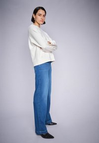 Lichtgrijze sweatshirt met verlaagde schouders, gecombineerd met een wijde spijkerbroek in blauw denim. Model draagt zwarte pumps met spitse neuzen. Eenvoudige achtergrond.