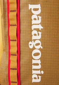 Bruine tas met textuur, featuring het witte "patagonia" logo, een rode accentstreep en een gele sectie met geweven details.