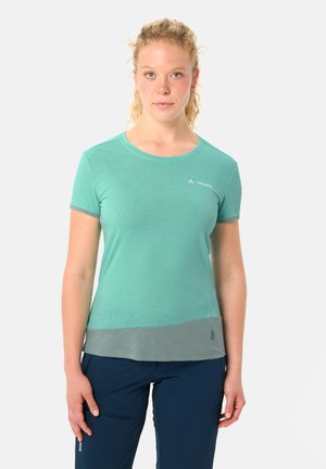 Jeune femme aux cheveux bouclés portant un t-shirt manches courtes turquoise et gris, et un pantalon bleu marine, debout devant un fond uni.