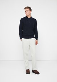 Granatowy sweter polo w splot kablowy z kołnierzykiem, w połączeniu z jasnoszarymi spodniami i brązowymi butami. Prosty design, gładka tekstura i dopasowany krój.