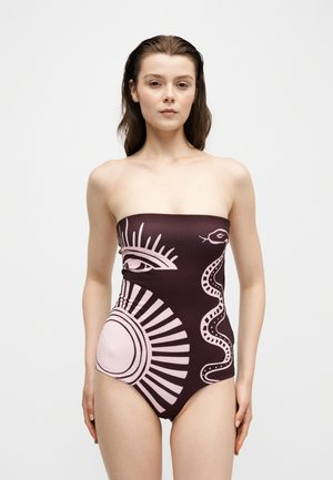 Femme portant un maillot de bain sombre sans bretelles avec de grandes illustrations rose clair d'œil, d'éclat de soleil et de serpent, debout devant un fond uni.