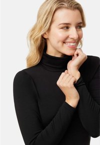 Top negro de cuello alto con mangas largas, textura suave, diseño ajustado, tejido suave. Las manos del modelo sostienen el escote, mostrando el estilo de la prenda.