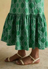 Grünes gemustertes Midikleid mit einem rüschengeschmückten Saum und einem geometrischen Design, kombiniert mit beigen flachen Sandalen mit dünnen Riemen und einer Schnallenapplikation.