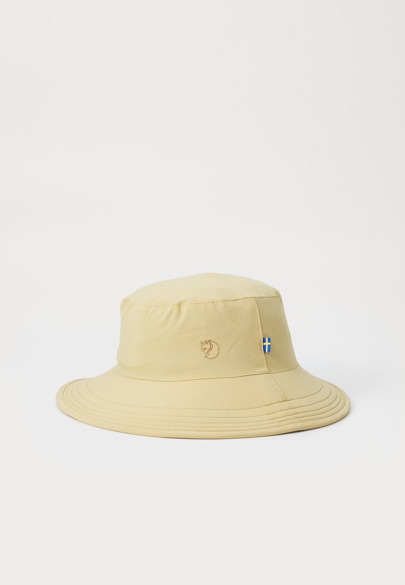 Beige bøttehatt laget av myk stoff med et sydd logo og en liten svensk flaggdetalj. Har en bred, buet skjerm og plisserte detaljer.