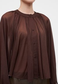 Blouse marron transparente à manches longues avec boutons recouverts de tissu et col froncé, portée par une personne des épaules à la taille.