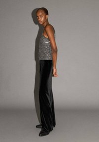 Strieborný flitrový top bez rukávov skombinovaný s čiernymi sametovými nohavicami s vysokým pásom a širokými nohavicami. Model stojí zboku, aby predviedol strih a textúru oblečenia.