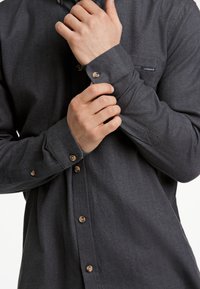 Camicia a bottoni a spina di pesce grigia scura con colletto a punta, chiusure con bottoni marroni e maniche lunghe. Presenta una piccola tasca sul petto.