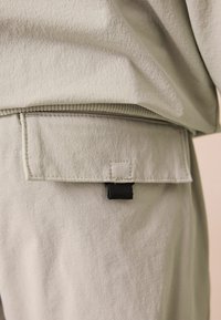 Vêtement gris clair en tissu texturé, doté d'un rabat de poche arrière et d'un accent en sangle noire pour une fonctionnalité accrue.