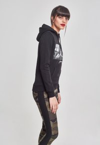 Sudadera negra con un bolsillo frontal y una impresión fotográfica. Combinada con leggings de camuflaje que cuentan con una franja lateral negra para dar contraste.