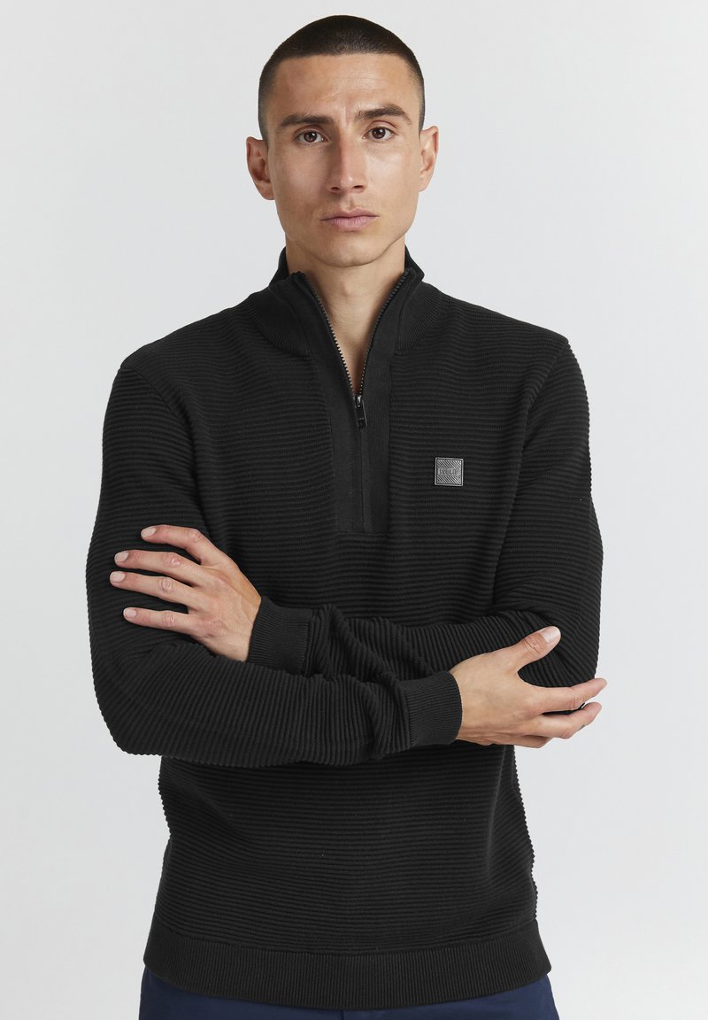 Schwarzer Reißverschluss-Pullover aus strukturiertem Rippstoff, Stehkragen und einem kleinen Logo-Patch auf der Brust. Lange Ärmel und enganliegende Bündchen.
