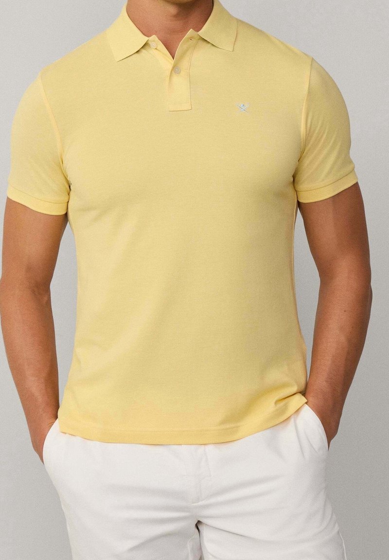 Homme portant un polo à manches courtes jaune pâle et un pantalon blanc, les mains dans les poches, devant un fond uni.