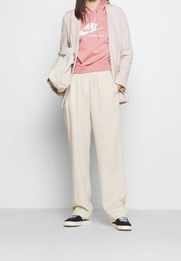 Sudadera de color rosa claro con logo blanco de Nike, pantalones beiges con cordones, y zapatillas negras con acentos blancos. Estilizado con un blazer claro.