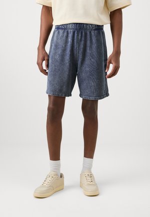 Shorts en denim bleu délavé avec une taille élastique, des poches latérales et des ourlets retroussés, associés à des baskets beige clair et des chaussettes blanches.