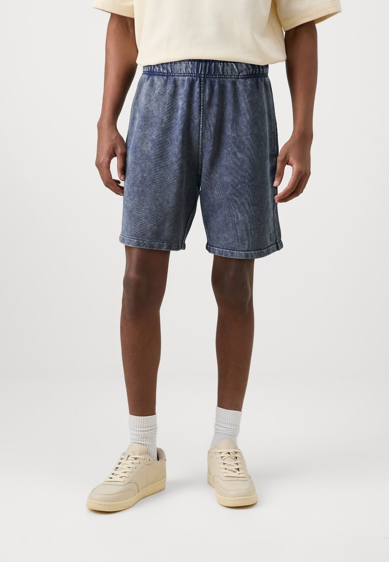 Shorts en denim bleu délavé avec une taille élastique, des poches latérales et des ourlets retroussés, associés à des baskets beige clair et des chaussettes blanches.
