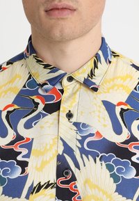 Chemise à manches courtes en tissu léger, présentant un motif coloré de grue et de nuages en jaune, bleu, rouge et noir, avec des boutons noirs.