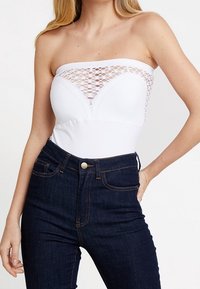 Valkoinen olkaimeton bodysuit, jossa on teksturoitu pitsimäinen muotoilu rinnassa, yhdistettynä korkeavyötäröisiin tummiin farkkuihin.