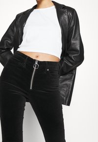 Černá kožená bunda přes bílý krátký top, doplněná vysokými černými manšestrovými kalhotami s stříbrným zipem a kroužkovým přívěskem.