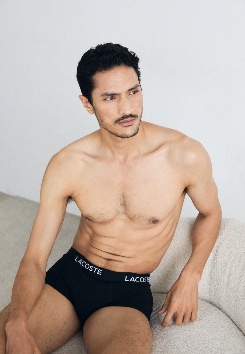 Black boxer briefs feature a white "LACOSTE" logo on the waistband. The model has a muscular build and is seated on a light-colored couch.