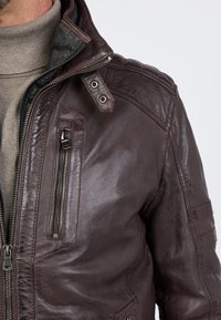 Bruine leren jas met een ritszak, gestructureerde kraag en doorgestikte details op de schouders. Voorzien van zilveren hardware-accenten.