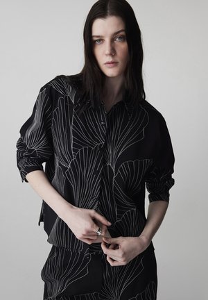 Jonge vrouw met lang donker haar, gekleed in een zwart outfit met witte abstracte lijnpatronen, die haar mouw rechttrekt tegen een effen achtergrond.