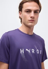 Νέος άνδρας που φοράει μωβ κοντομάνικο πουκάμισο με το λογότυπο "HAROX" και το σήμα της Puma στο μπροστινό μέρος, κοιτάζοντας προς τα εμπρός μπροστά σε απλό φόντο.