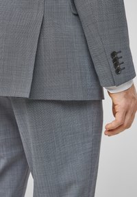 Lichtgrijs pak, met een gestructureerde stof en een klein ruitpatroon. Bevat zwarte knopen op de manchetten en een recht gesneden jasje en broek.