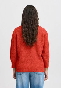 Pull en tricot rouge avec des manches trois-quarts, une finition texturée et un décolleté large ; associé à un jean en denim bleu clair.