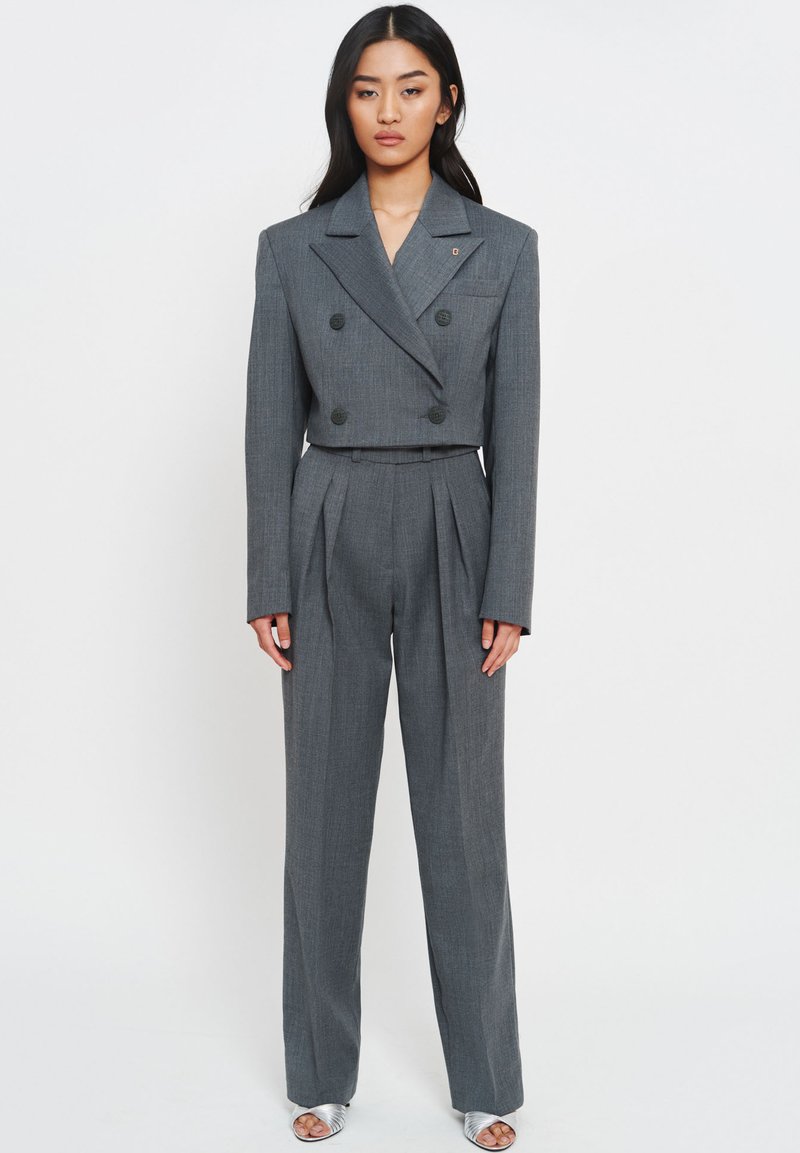Grijs dubbelbörstig pak van gestructureerde stof, met een verkorte jasje, brede broek met plooien en zwarte knopen. Zilveren sandalen.
