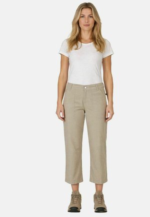 Frau mit langen blonden Haaren, die ein weißes T-Shirt mit kurzen Ärmeln, beige kurze Hosen und beige Wanderschuhe trägt, steht vor einem schlichten Hintergrund.