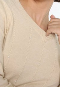 Beżowy sweter dziany z dekoltem w serek, z teksturowanymi diagonalnymi liniami i małymi perłowymi akcentami wzdłuż wzoru. Miękka tkanina i dopasowany krój.