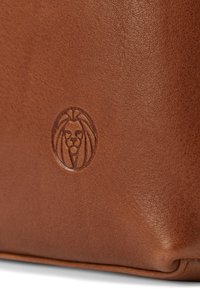 Ruskea nahkabagi, jonka pinnassa on teksturoitu pinta ja kohokuvioitu leijonan logo. Muoto on suorakulmainen pyöristettyine reunoineen, mikä korostaa käsityön laatua.