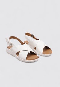 Sandalias de cuero blancas con correas cruzadas, plantilla marrón, una correa ajustable en el tobillo y una suela baja de goma blanca.