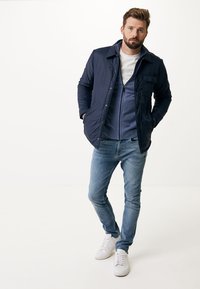 Námornícky prošívaný kabát s na knoflíky, kapsou na hrudi, nošený přes světle modrou mikinu na zip, kombinovaný se světle modrými džíny a bílými teniskami.