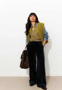 Gilet en polaire vert olive porté sur une chemise en denim, un haut à motifs paisley et un pantalon large en velours noir. Sac fourre-tout à carreaux marron et chaussures sombres.