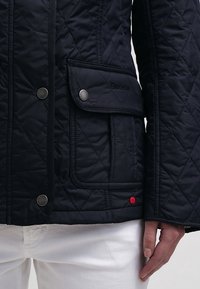 Gros plan d'une veste matelassée noire avec poche à rabat et boutons-pression métalliques, portée avec un pantalon blanc, mettant en avant la texture et les détails de couture.
