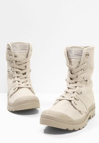 Bottes montantes en toile beige avec bout rond, semelle en caoutchouc, et œillets métalliques. Caractéristiques comprenant un col rembourré et un tissu texturé pour une meilleure adhérence.